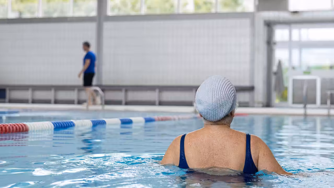 Nageuse senior vue de dos dans un bassin intérieur, près d’une ligne d’eau et sous la surveillance d’un maître-nageur