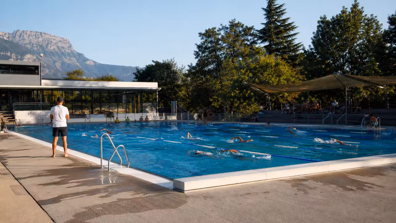 Bassin municipal extérieur fréquenté en été, avec nageurs, plage ombragée et montagnes autour de Grenoble