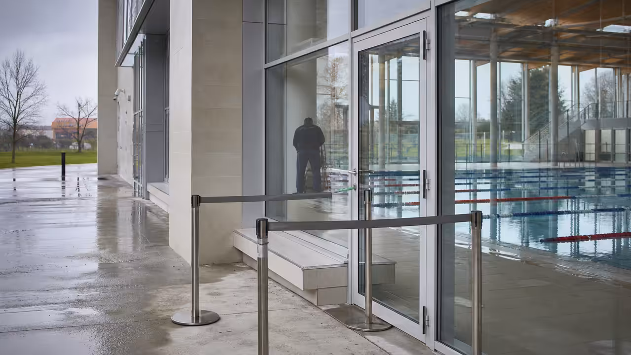 Entrée fermée d’un centre aquatique municipal, avec bassins visibles derrière de grandes baies vitrées