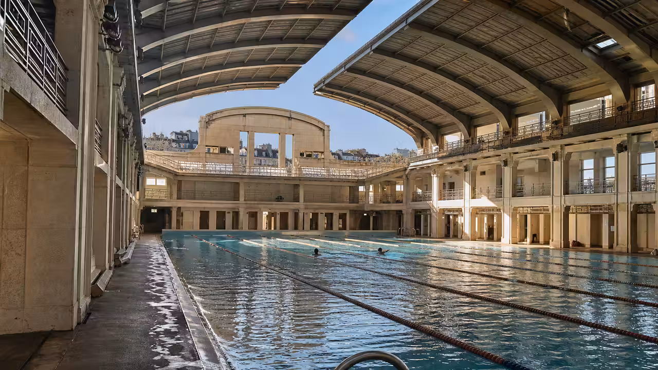 Bassin de natation couvert baigné de lumière naturelle sous le toit ouvrant d’une piscine parisienne