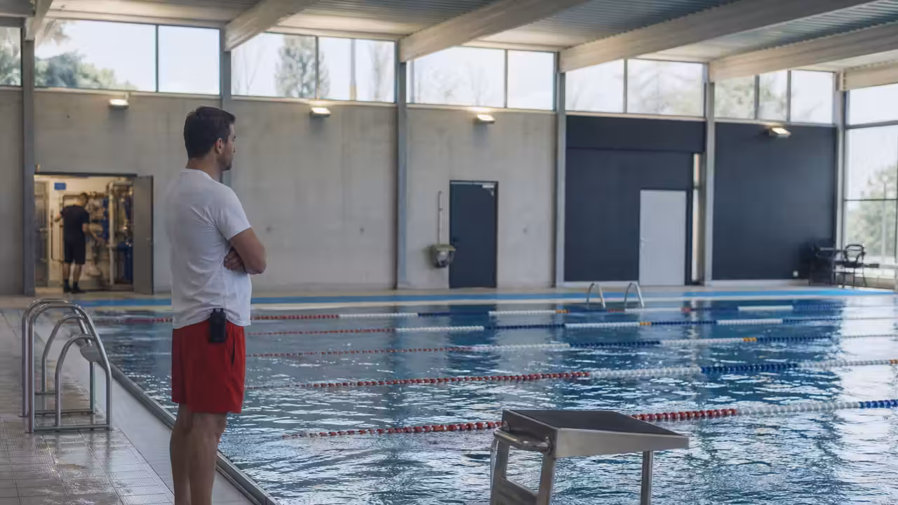 Maître-nageur observant un bassin couvert pendant qu’un agent technique contrôle les installations en arrière-plan.