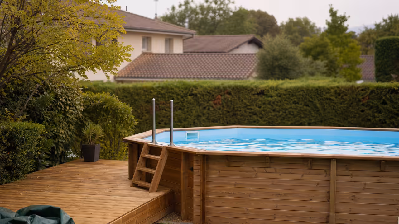 Piscine hors-sol en bois installée dans un jardin, avec échelle relevée et terrasse en arrière-plan