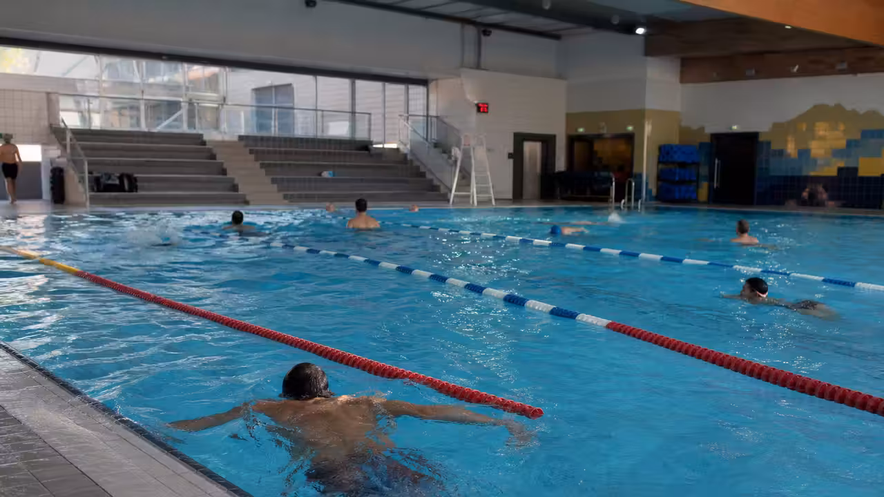 Bassin intérieur d’une piscine municipale avec lignes d’eau, baigneurs au loin et maître-nageur en surveillance
