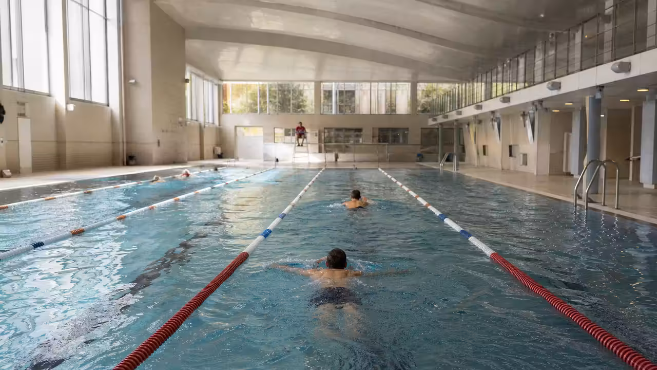 Nageurs dans un bassin couvert parisien lumineux, avec lignes d’eau et maître-nageur au loin