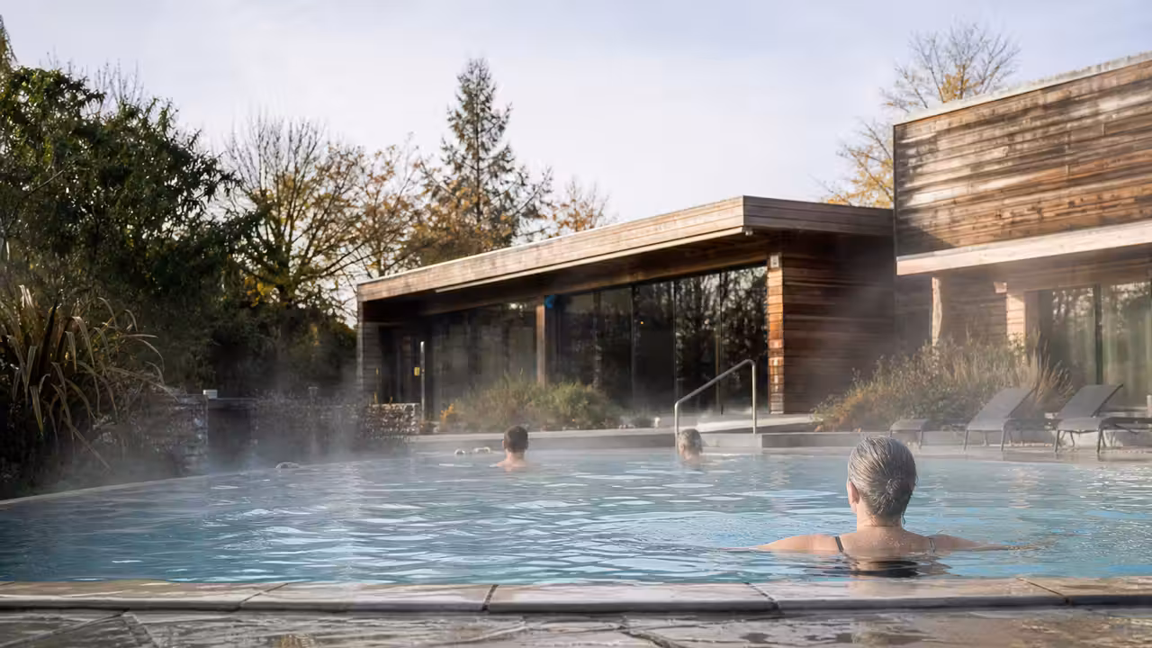 Bassin extérieur chauffé d’un centre aquatique, bordé de végétation, utilisé par des nageurs en saison fraîche