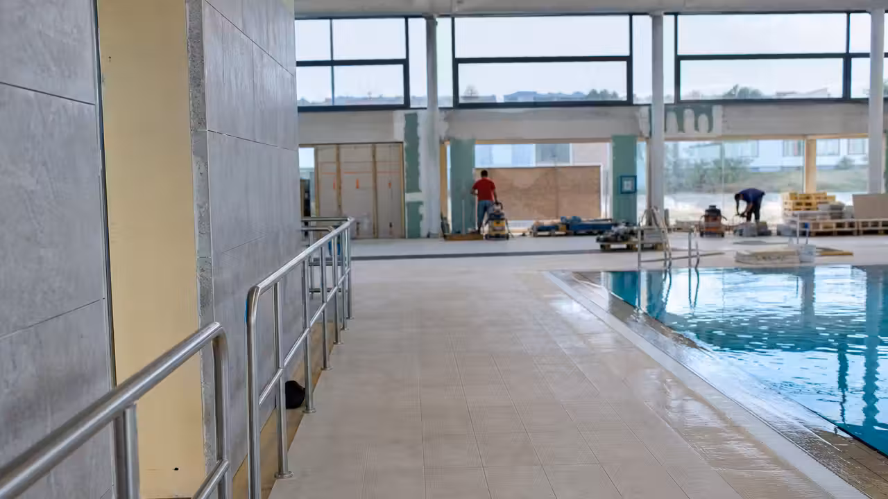 Hall lumineux d’une piscine municipale en rénovation, avec accès de plain-pied et vestiaires au second plan