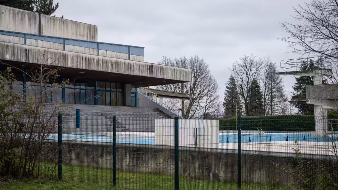 Ancien complexe aquatique municipal fermé, clôturé, avec bassin vide et bâtiments techniques en arrière-plan