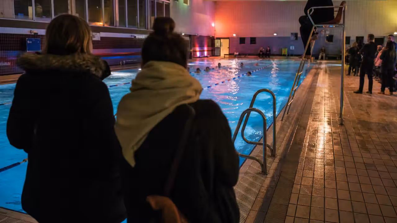 Bassin intérieur animé en soirée, avec éclairage coloré discret et baigneurs vus de dos à distance