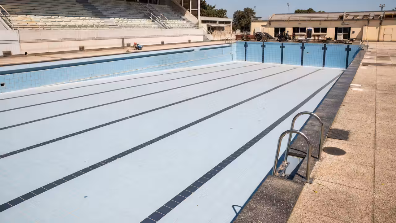 Bassin extérieur municipal temporairement vide sous un soleil intense, avec lignes d’eau et gradins déserts