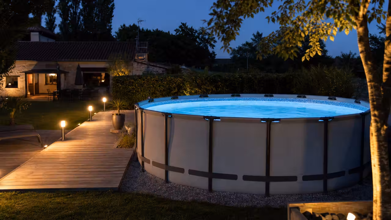 Piscine hors sol ronde éclairée en bleu au crépuscule sur une terrasse de jardin aménagée