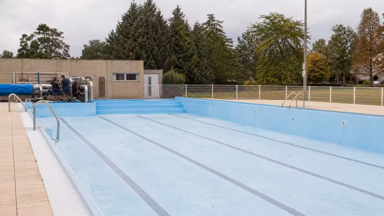 Bassin extérieur municipal vide en fin d’été, avec lignes d’eau retirées et équipes techniques au loin