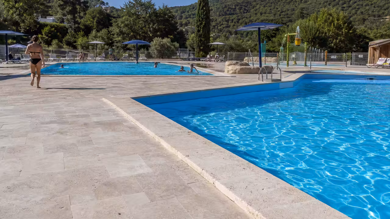 Bassin extérieur d’une piscine municipale au soleil, avec plage minérale et collines verdoyantes en arrière-plan