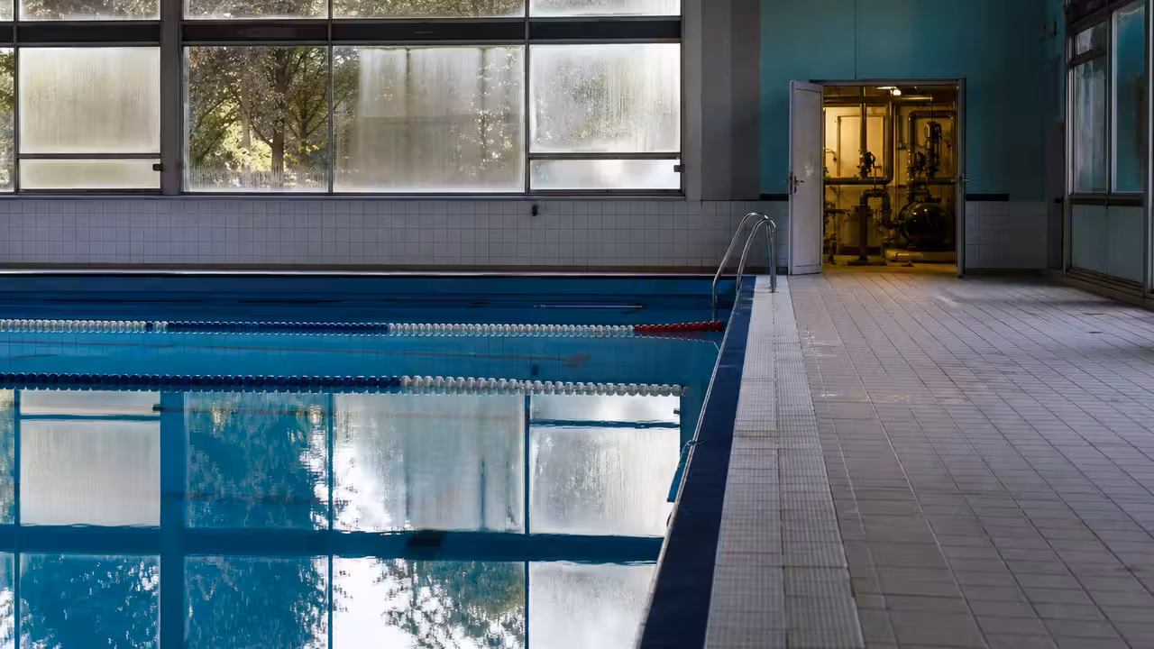 Bassin municipal intérieur vide avec lignes d’eau, baies vitrées et équipements techniques visibles en arrière-plan