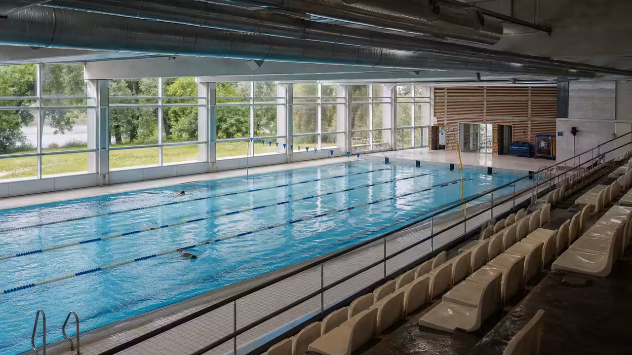 Bassin municipal couvert vu depuis les gradins, avec lignes d’eau, baies vitrées et équipements techniques discrets