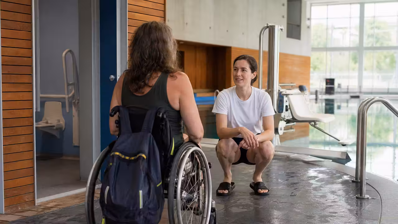 Entrée d’un centre aquatique municipal avec un adulte en fauteuil roulant accueilli près d’un bassin.