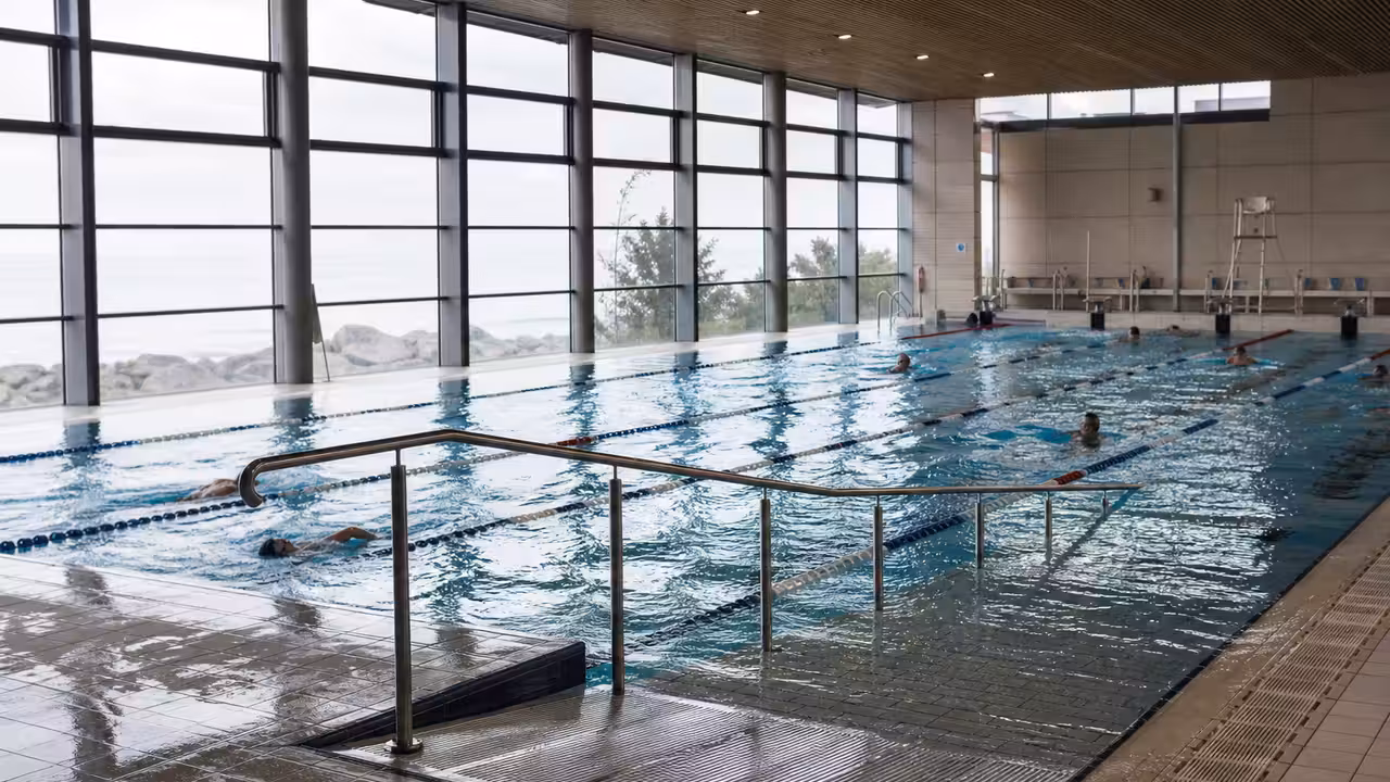 Nageurs dans un bassin municipal lumineux avec ligne d’eau, rampe d’accès et grandes baies vitrées