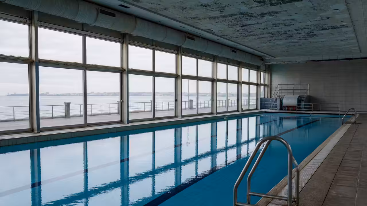 Bassin intérieur vide d’un centre aquatique municipal, avec baies vitrées donnant sur le front de mer