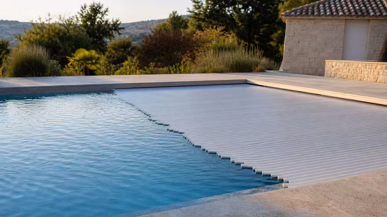 Volet roulant à lames blanches partiellement ouvert sur une piscine moderne bordée d’une terrasse minérale