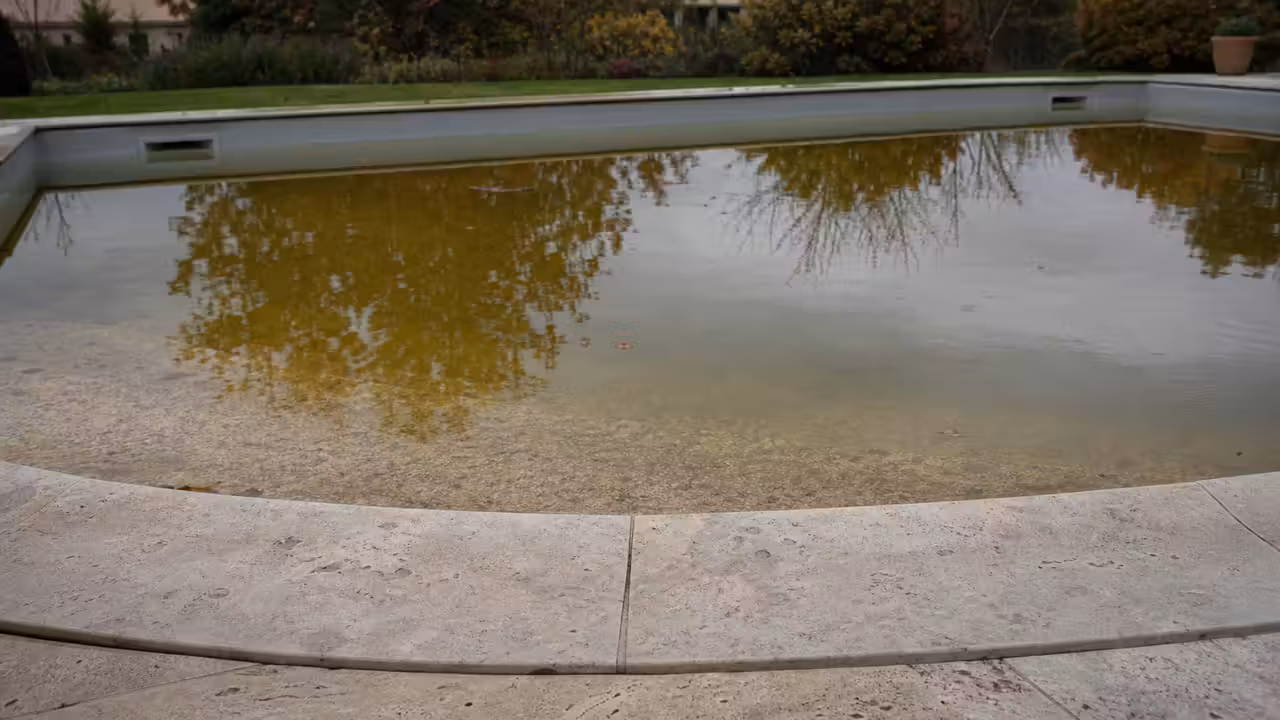 Piscine enterrée à l'eau teintée de brun ambré sous un ciel d'automne couvert, margelles en pierre claire
