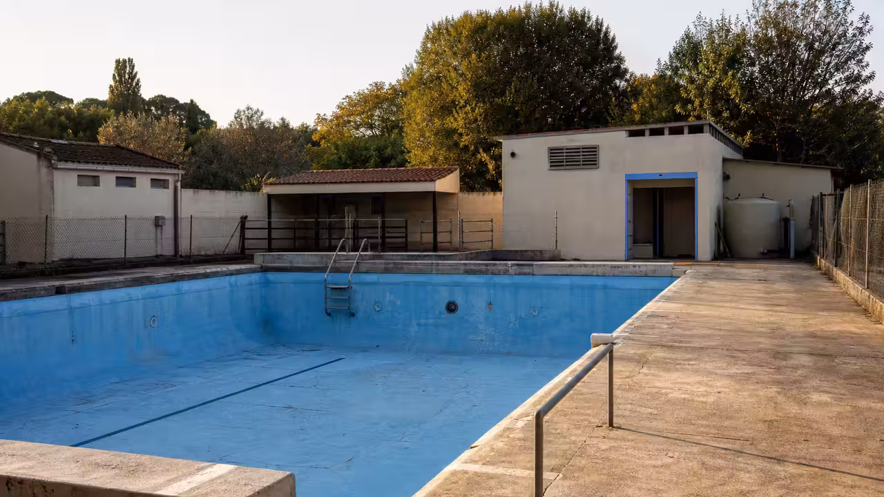 Bassin municipal extérieur vide avec équipements techniques vieillissants et bâtiment communal en arrière-plan