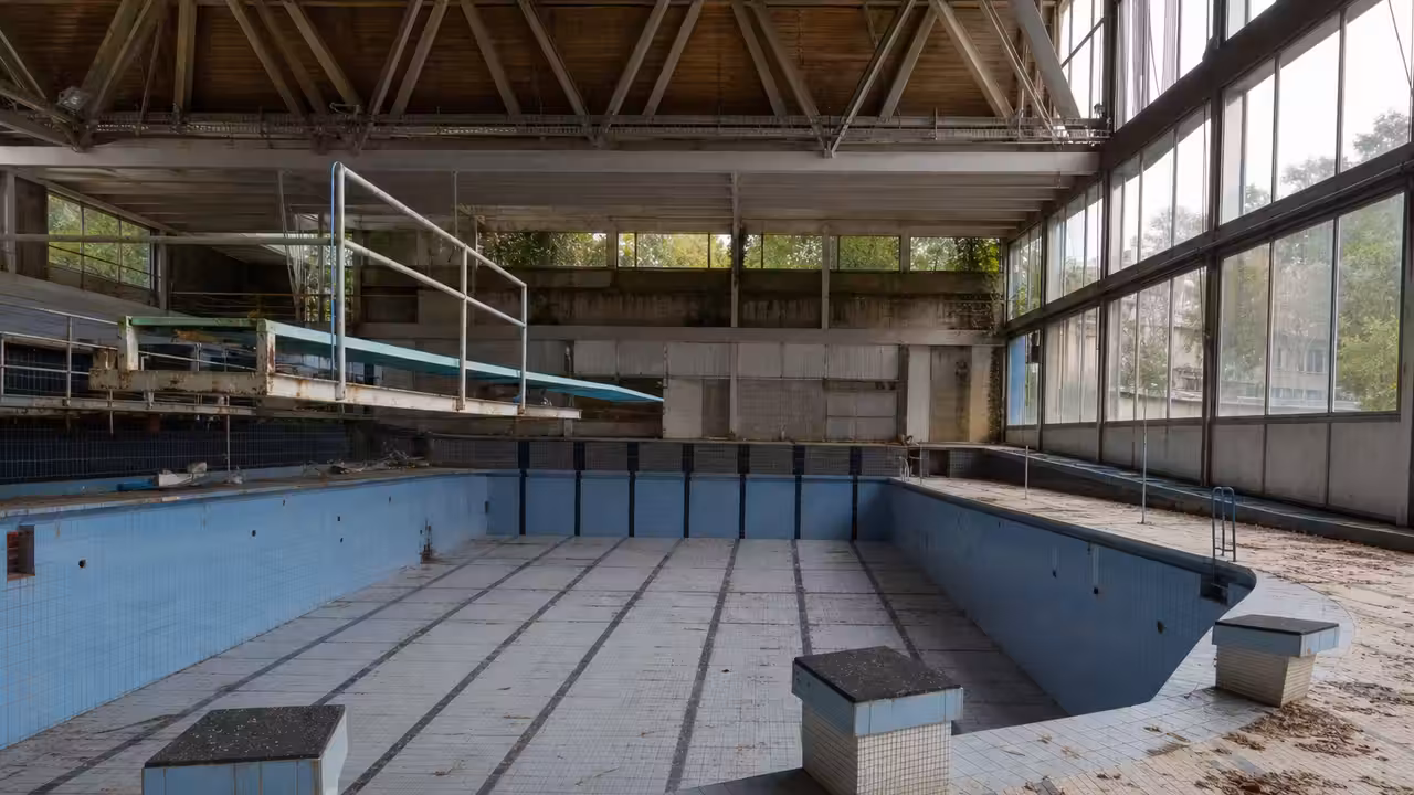 Ancien bassin municipal vide aux carrelages usés, éclairé par la lumière naturelle venant des baies vitrées