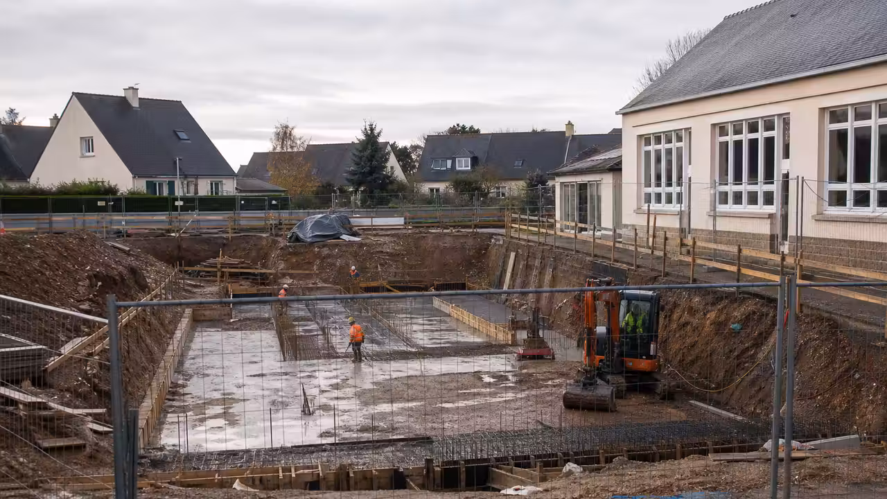 Bassin d’apprentissage en chantier près d’une école, avec zone de travaux clôturée et engin de terrassement