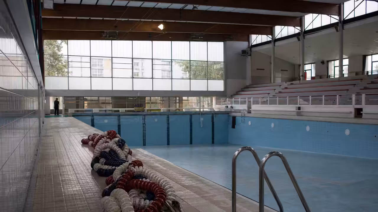 Bassin municipal couvert et vide, avec lignes d’eau retirées et lumière naturelle sur les gradins