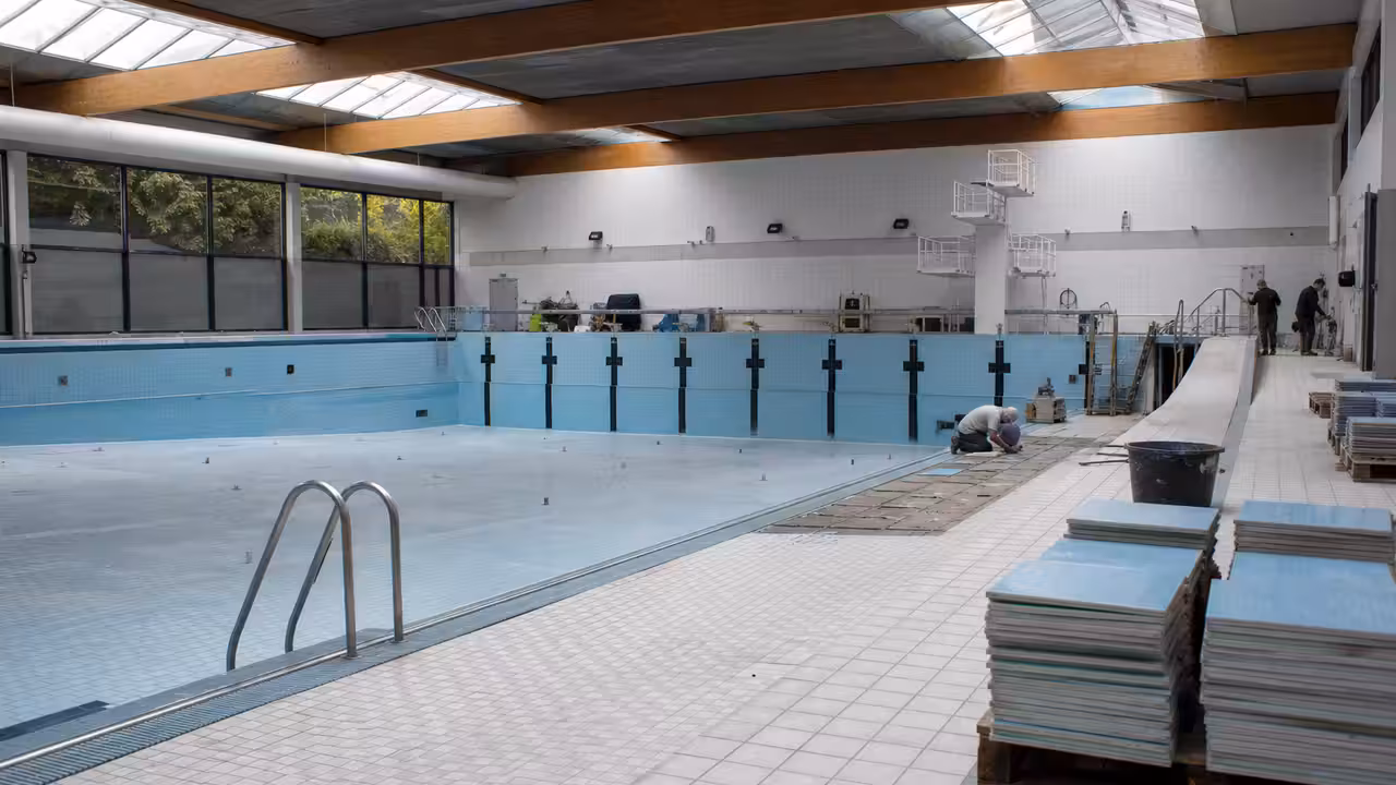 Bassin municipal vide en travaux, avec carrelage déposé et équipements techniques visibles sur les plages