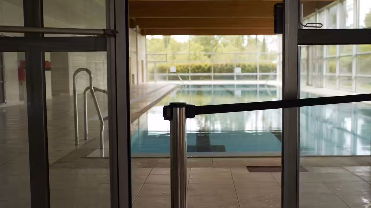 Entrée vitrée d’un centre aquatique public fermée, avec un bassin intérieur vide visible à l’arrière-plan