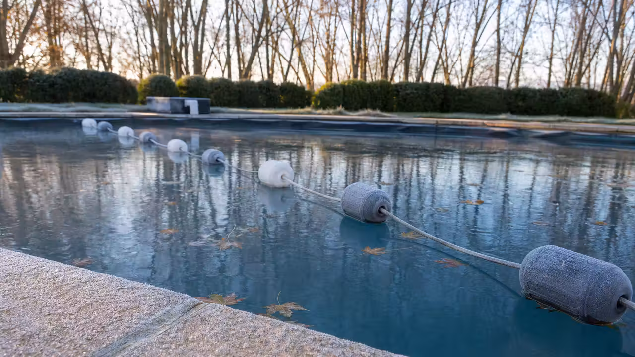 Ligne de flotteurs d'hivernage tendue en diagonale sur une piscine au petit matin, margelle couverte de givre