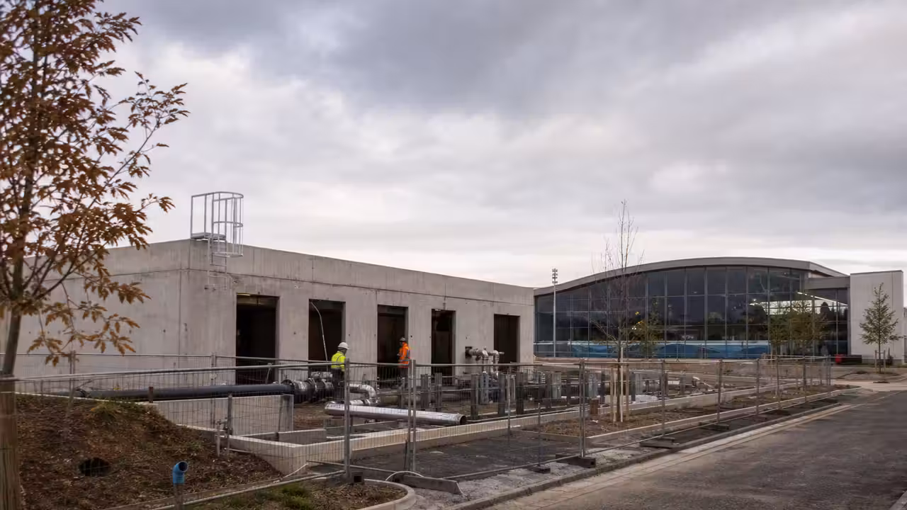 Bâtiment technique contemporain en construction à côté d’un centre aquatique municipal, avec réseaux visibles