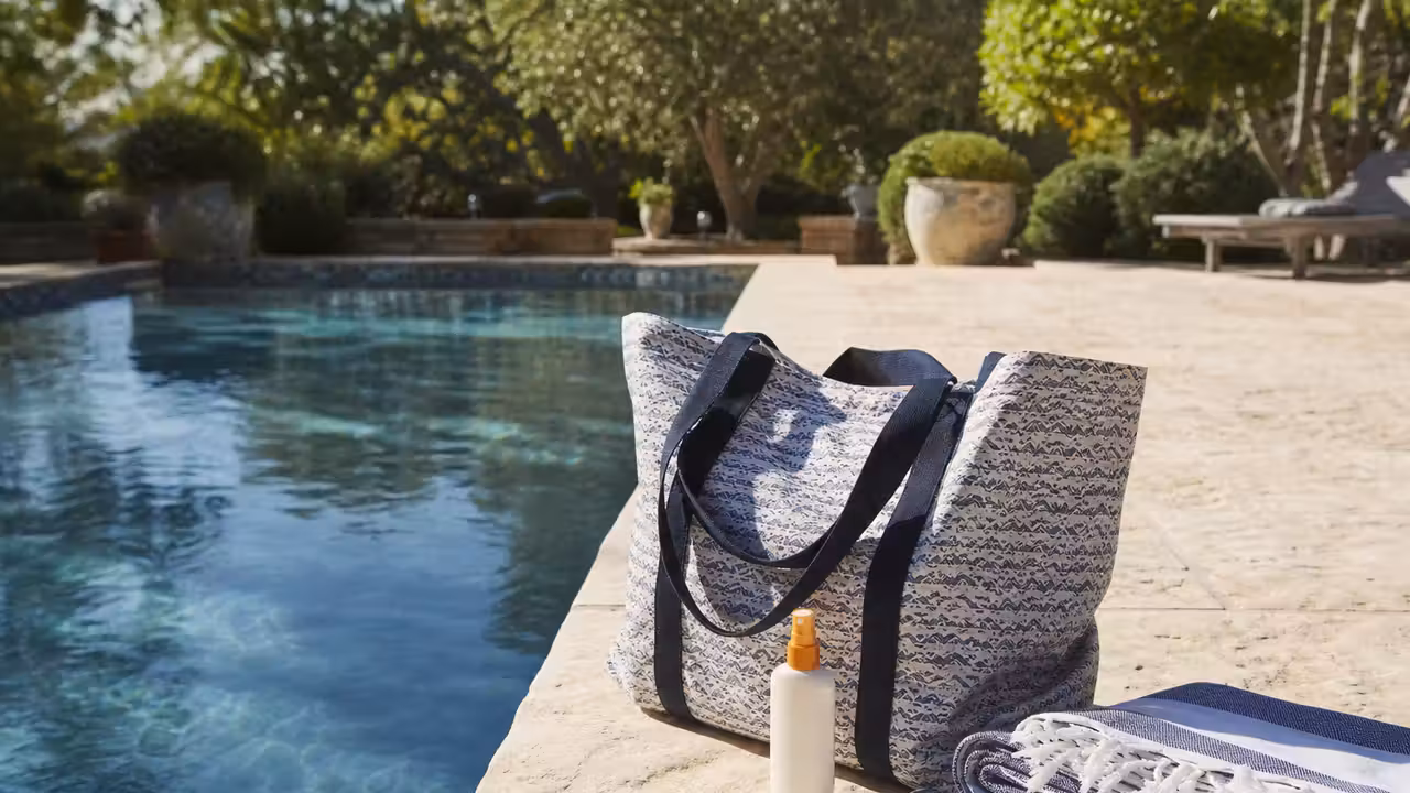 Grand sac de plage fermé posé sur une terrasse près d’une piscine, avec serviettes et pochette étanche.