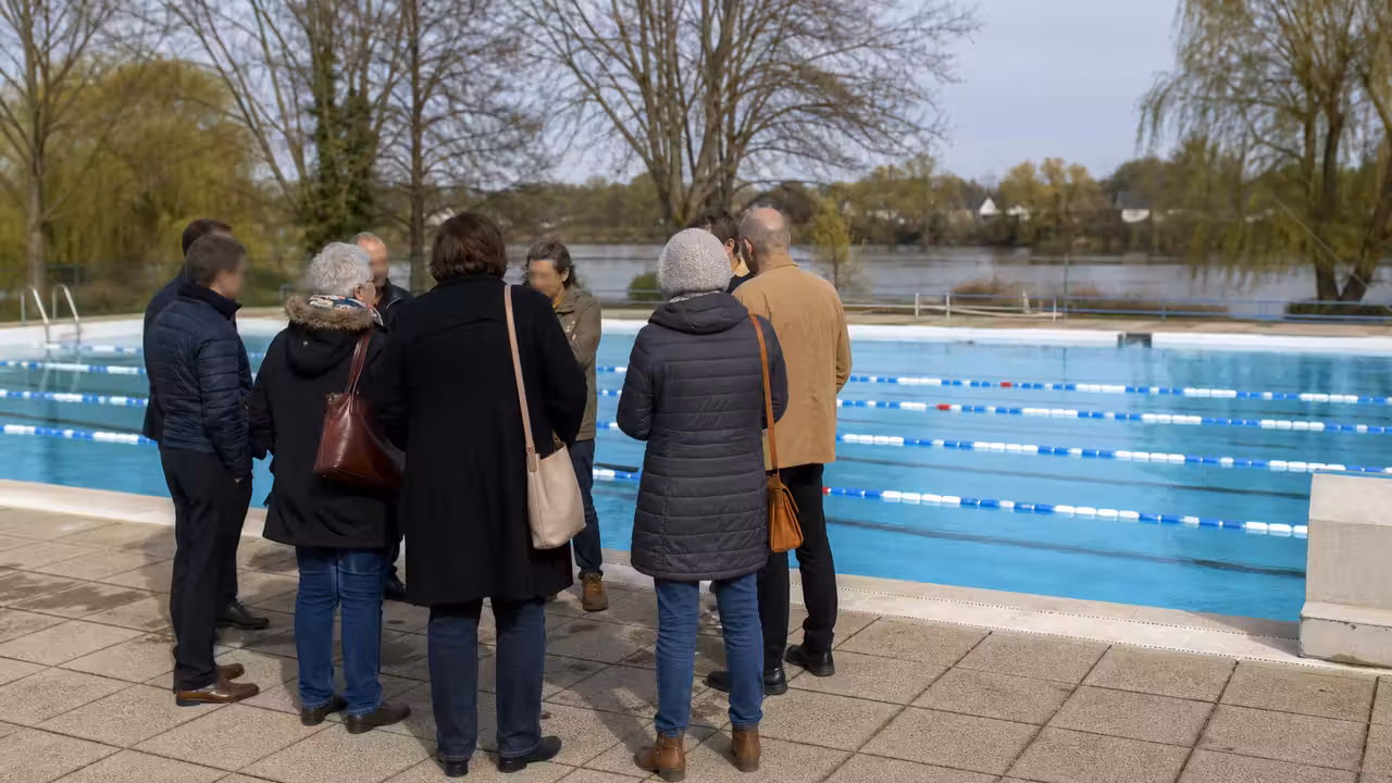 Bassin municipal extérieur avec lignes de nage, gradins sobres et habitants réunis à distance sur la plage