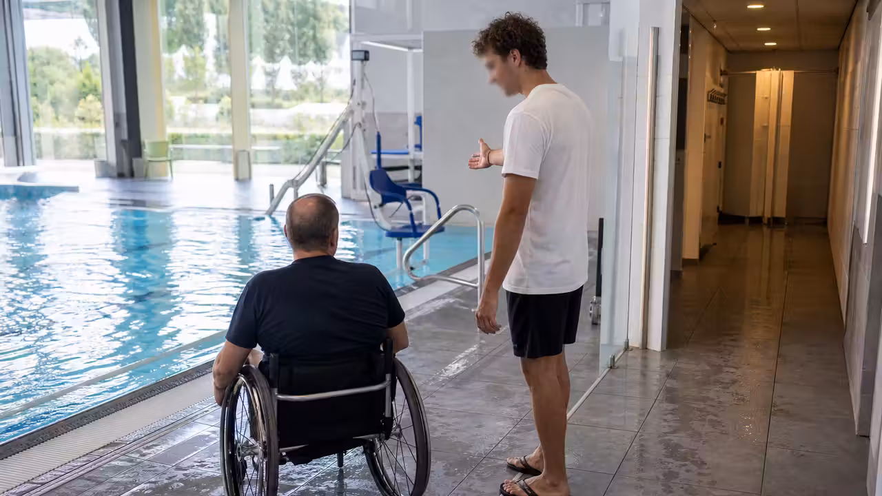 Personne en fauteuil accompagnée par un maître-nageur au bord d’un bassin public accessible et lumineux