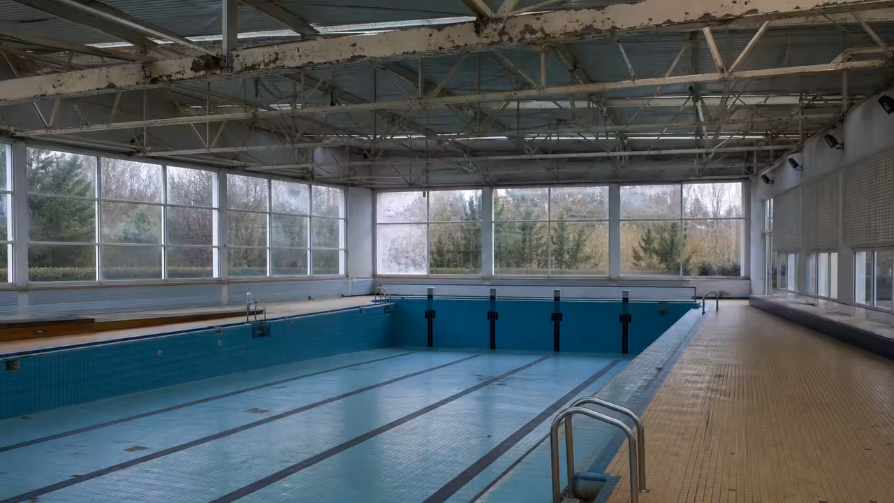 Intérieur d’une piscine municipale ancienne fermée, avec charpente métallique et bassin vide sous lumière naturelle.