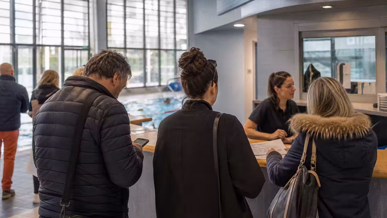 Usagers adultes consultent les créneaux de cours au comptoir d’accueil d’une piscine publique lumineuse.