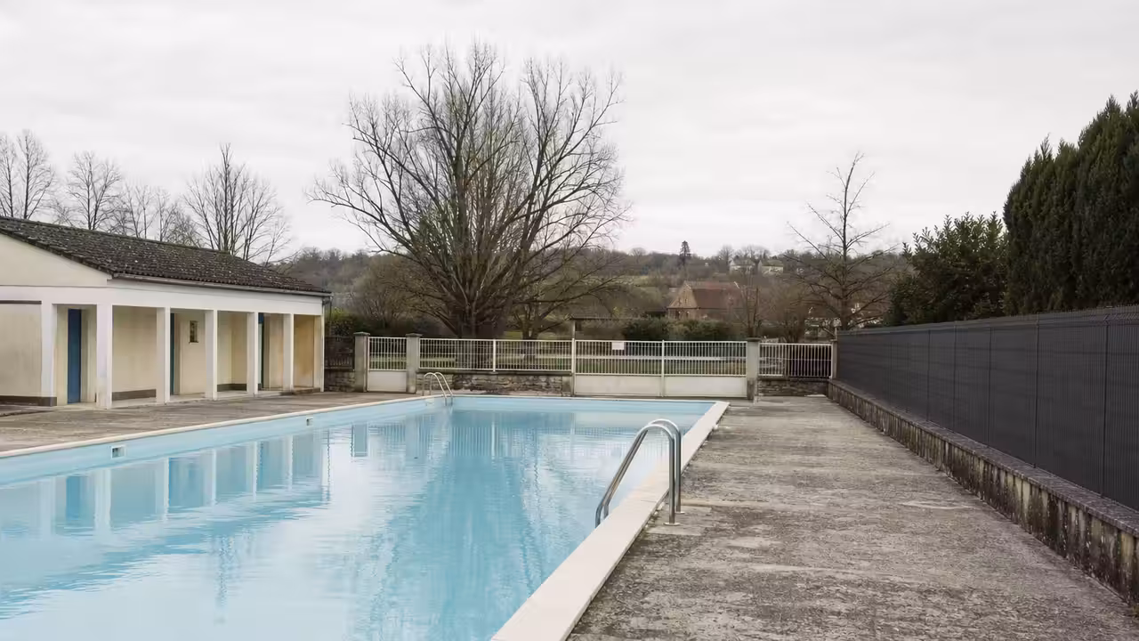 Bassin extérieur municipal vide, gradins simples et arbres d’hiver évoquant la fermeture d’une piscine locale