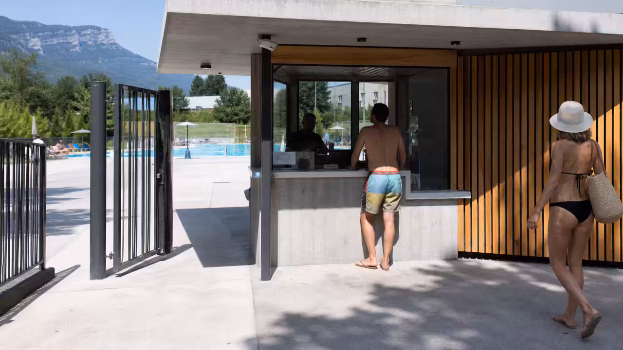 Entrée d’une piscine municipale avec portillon, bassin extérieur et agents d’accueil en arrière-plan