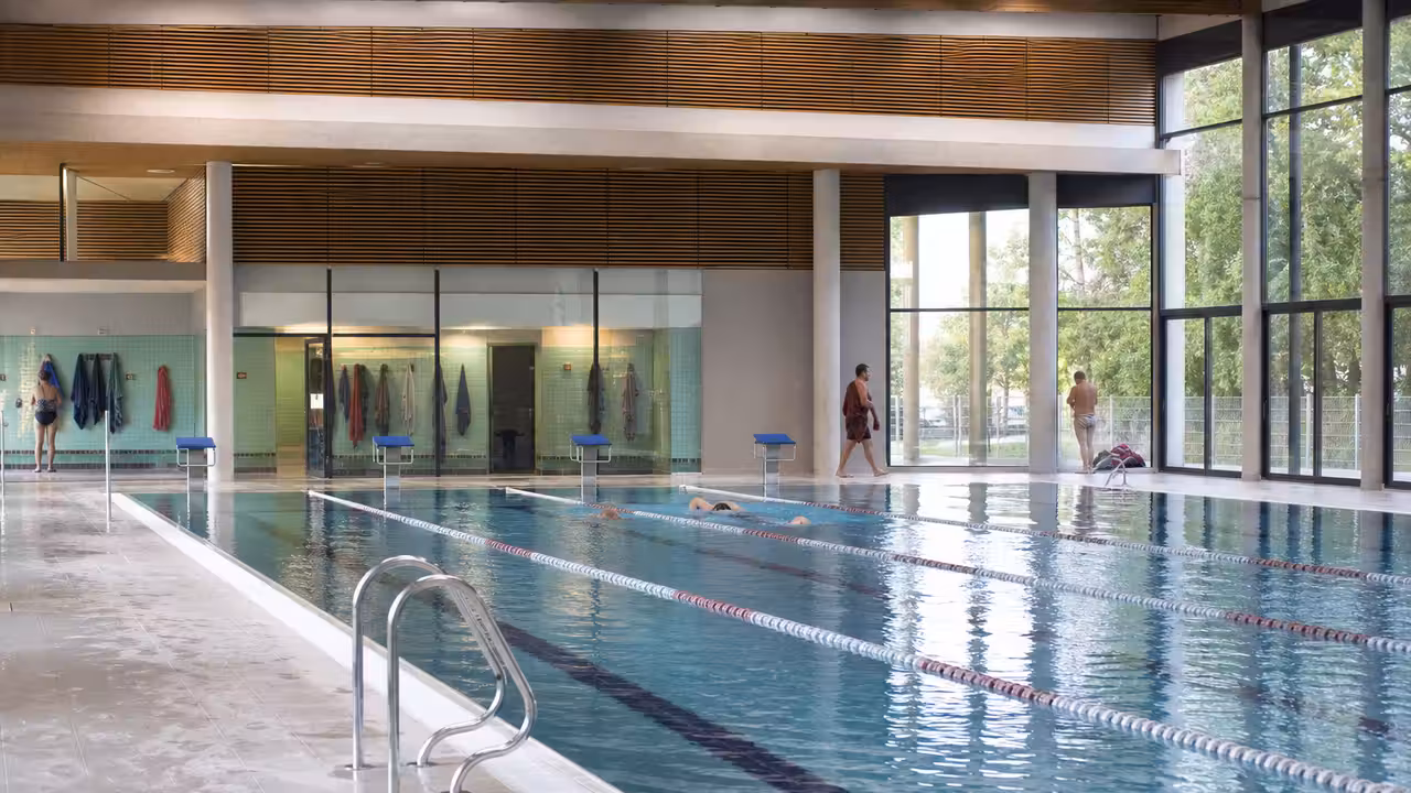 Bassin intérieur rénové avec lignes de nage, baigneurs en arrière-plan et grandes baies vitrées lumineuses