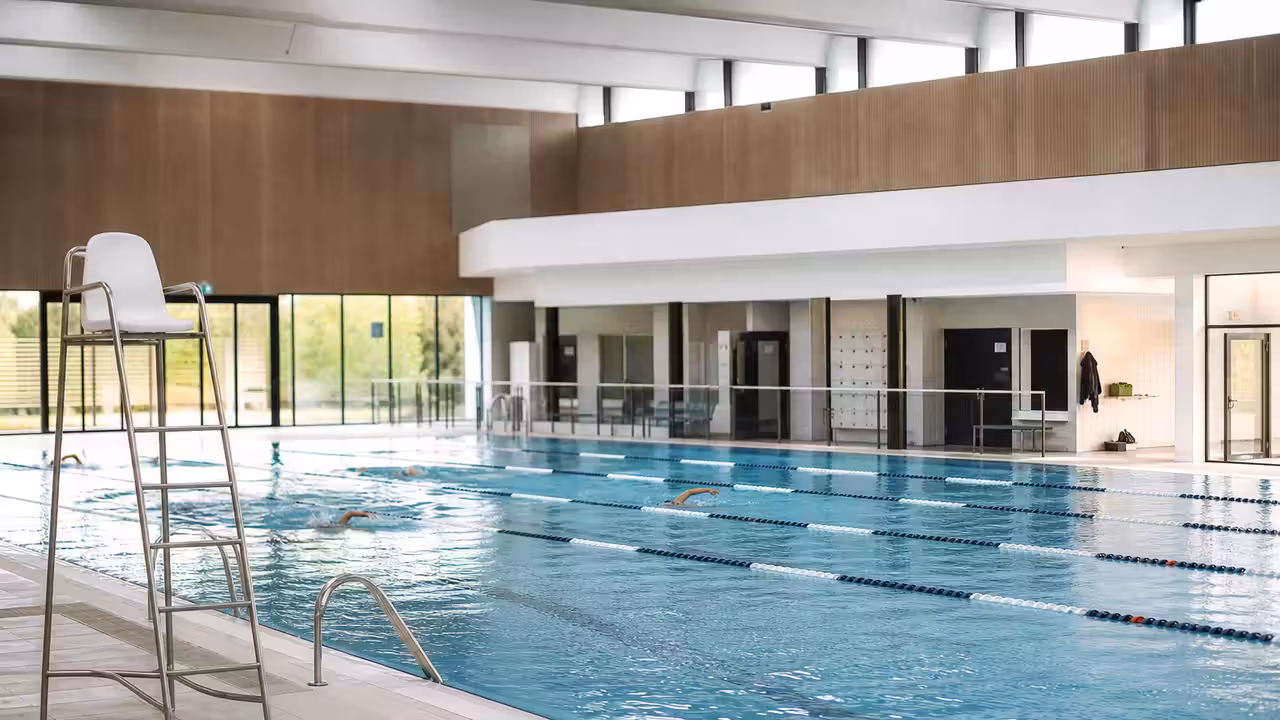 Bassin intérieur rénové d’une piscine municipale, avec lignes d’eau, vestiaires vitrés et baigneurs au loin