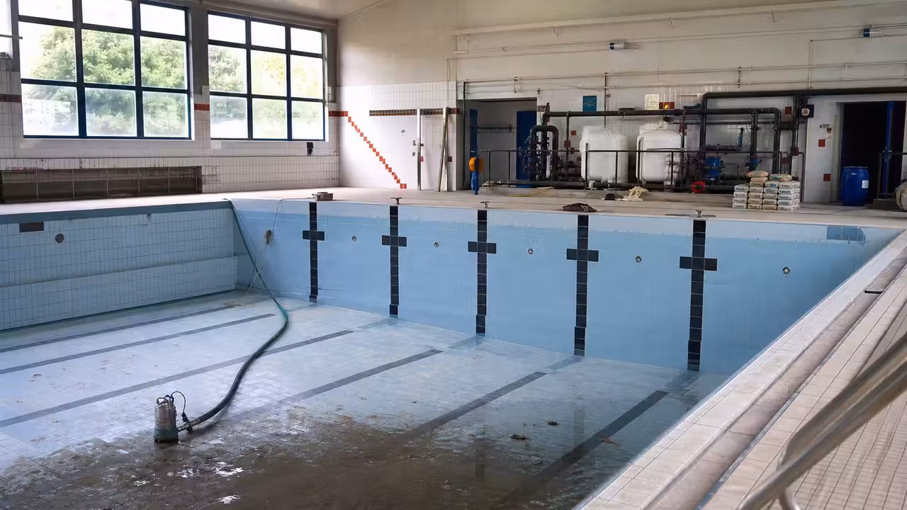 Bassin couvert municipal vide pendant une rénovation, avec réseaux techniques et baies vitrées en arrière-plan
