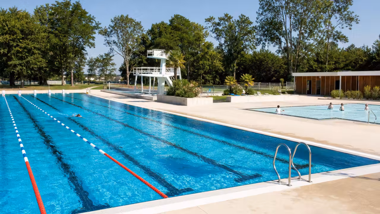 Bassins rénovés d’une piscine municipale avec plage minérale, espaces verts et nageurs au loin