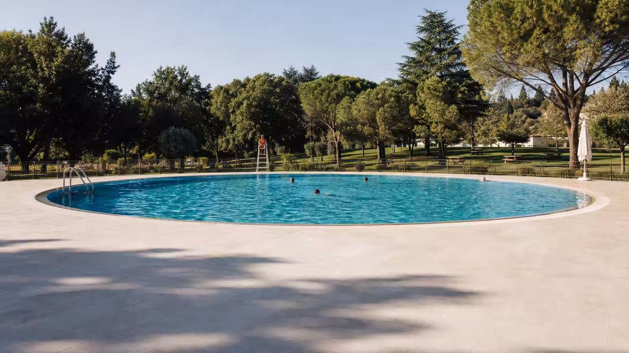 Bassin circulaire extérieur rénové, plages minérales et arbres autour d’une piscine publique estivale