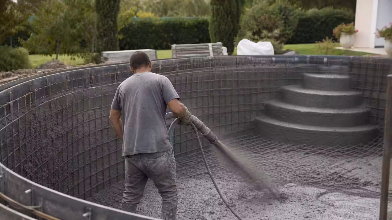 Projection de béton sur l’armature d’une piscine en construction, avec parois courbes et chantier préparé.