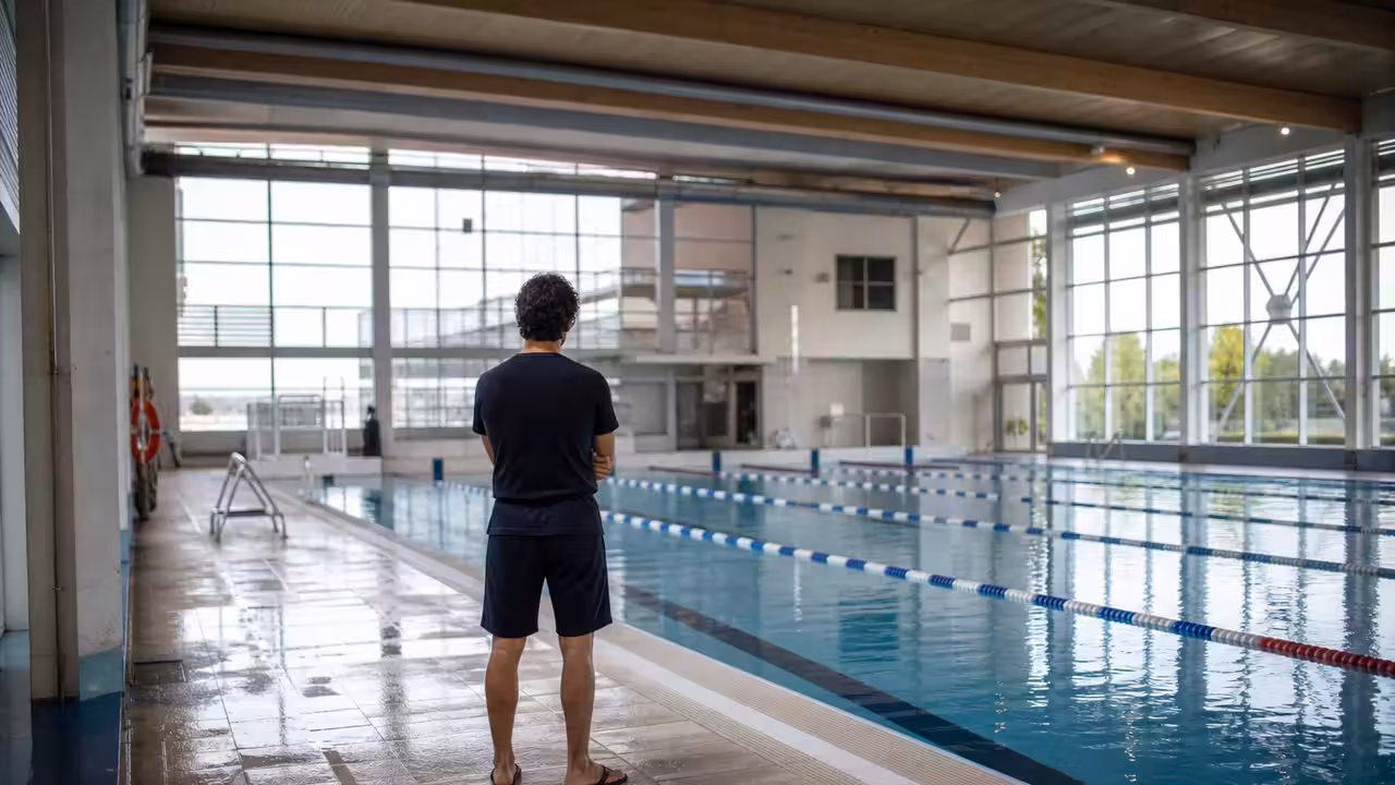 Bassin couvert de piscine publique avec lignes d’eau, gradins vides et maître-nageur observant depuis le bord