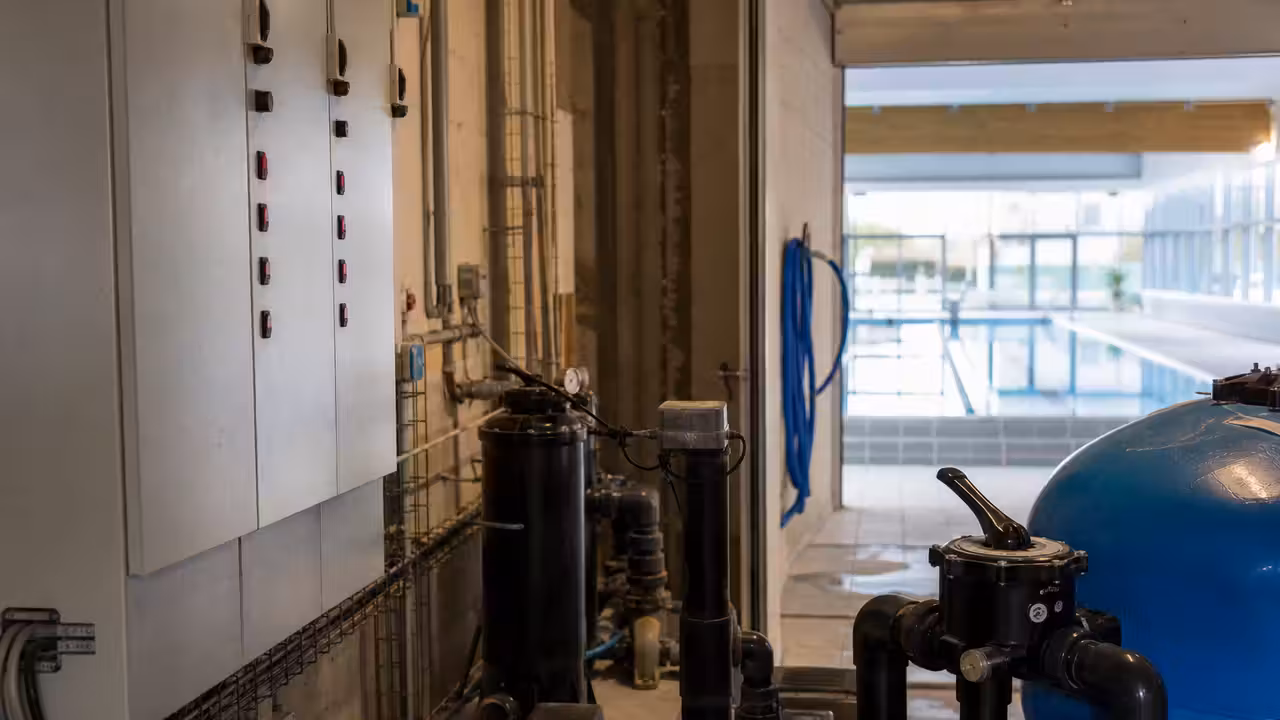Local technique d’une piscine municipale avec tableaux électriques et bassin vide en arrière-plan