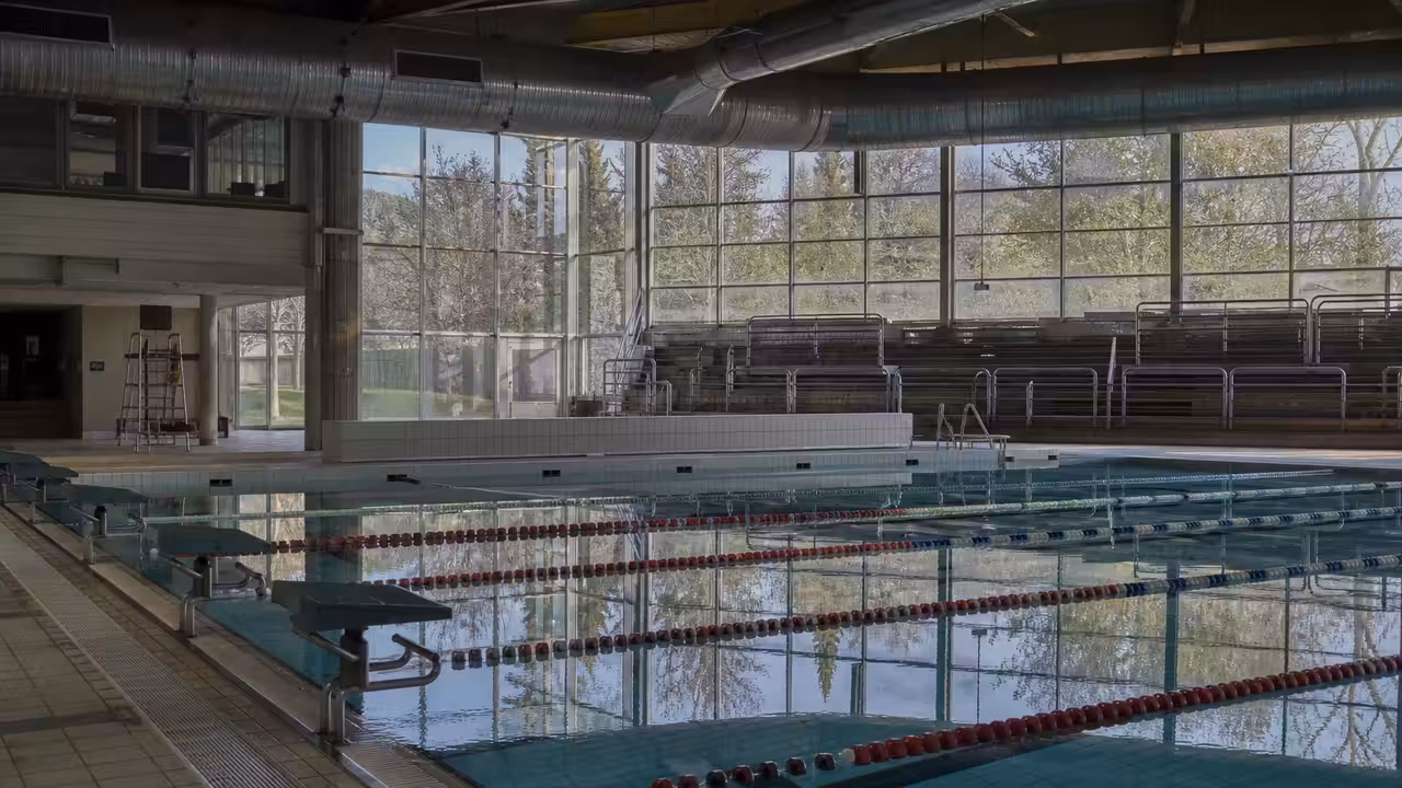 Bassin de piscine municipale vide au petit matin, avec lignes d’eau, gradins et baies vitrées en arrière-plan.