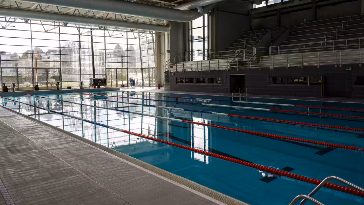 Bassin couvert municipal vide avec lignes d’eau, gradins sobres et équipements techniques en arrière-plan