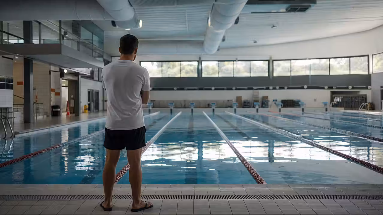 Maître-nageur de dos au bord d’un bassin couvert, sous les bouches de ventilation d’une piscine municipale.