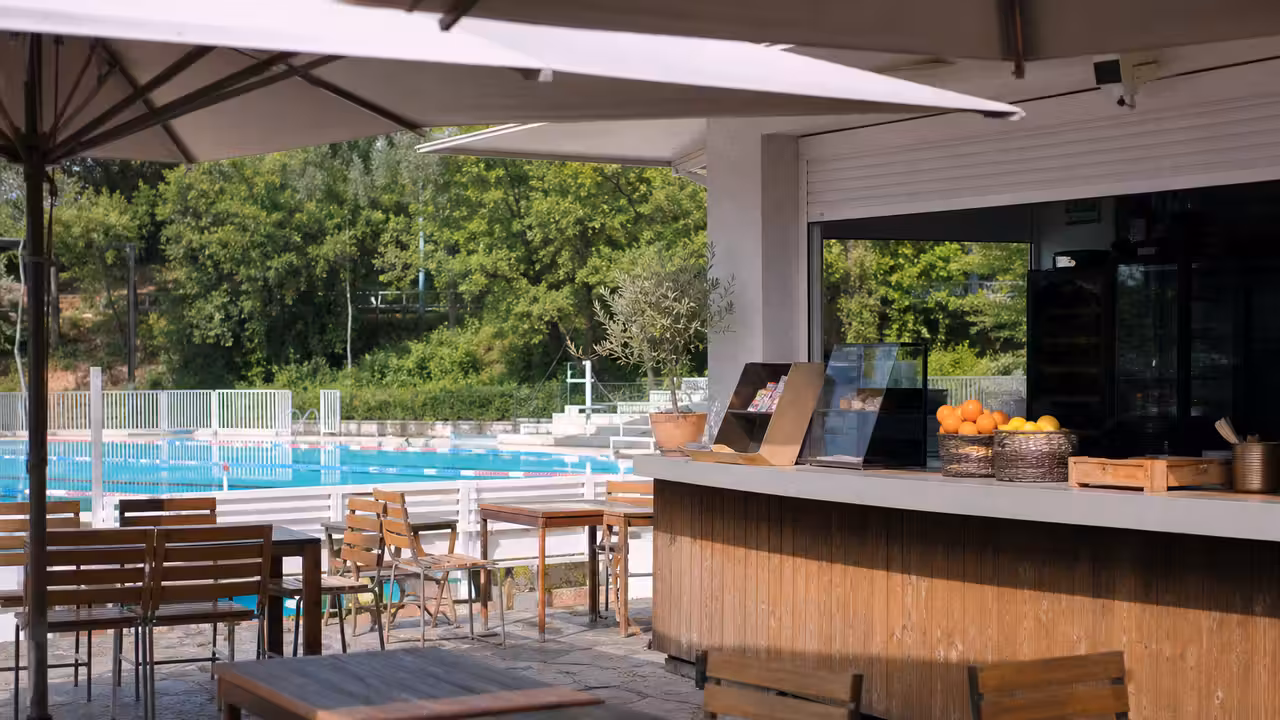 Terrasse de snack ombragée au bord d’une piscine publique, avec bassins et transats en arrière-plan